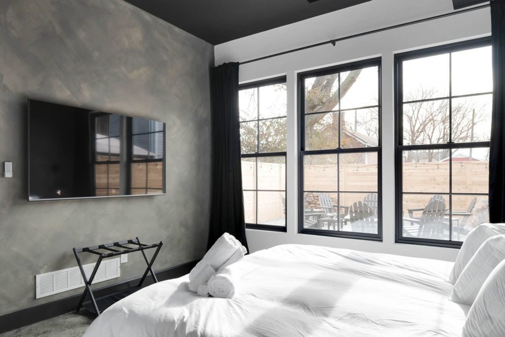 Louisville modern bedroom with minimalist white bed, black-framed windows, wall-mounted TV, and scenic tree views.