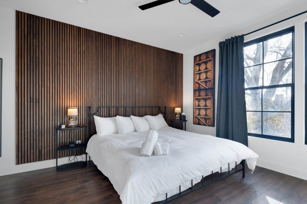 Louisville modern bedroom with white linens, wood-paneled accent wall, and large window, highlighting custom home design ideas.