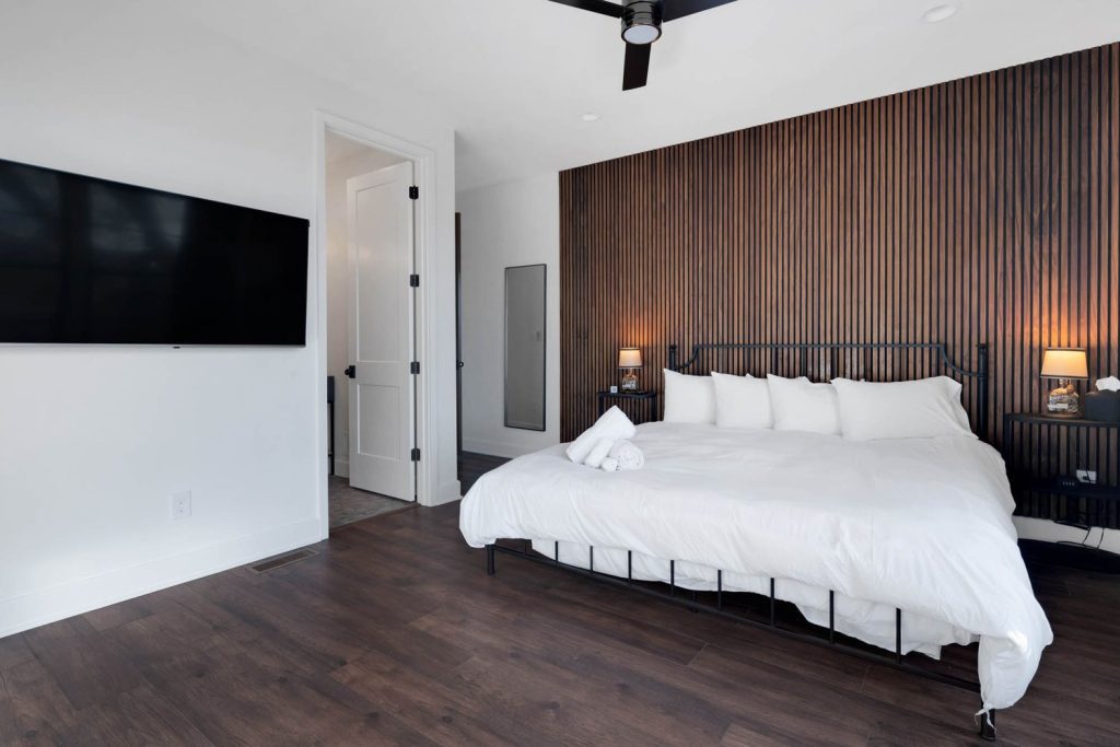 Louisville modern bedroom with white bed, wood accent wall, wall-mounted TV, and soft lighting by top custom home builders.