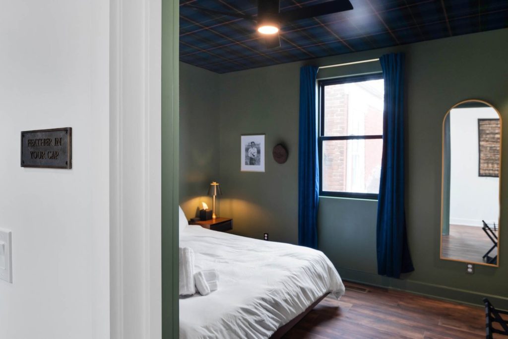 Cozy modern bedroom with white bedspread, deep green walls, blue curtains, and plaid ceiling. Warm lamp light creates a serene, inviting ambiance.