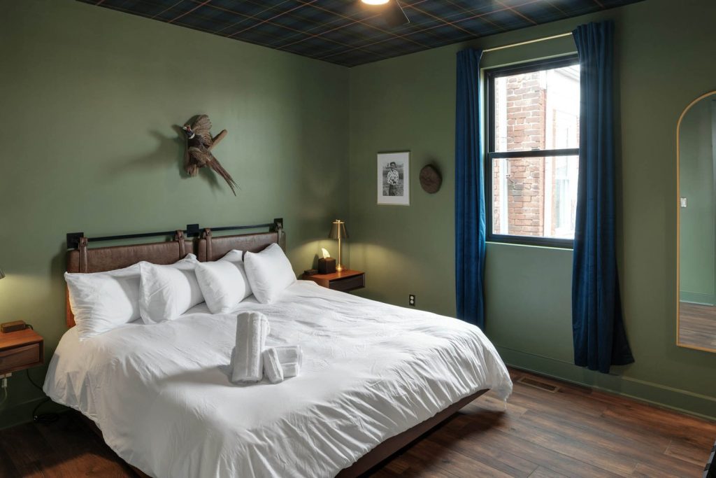 Cozy modern bedroom with green walls, white bed linens, gold lamp, blue curtains, wall art, and wooden floors. Calm, inviting ambiance.