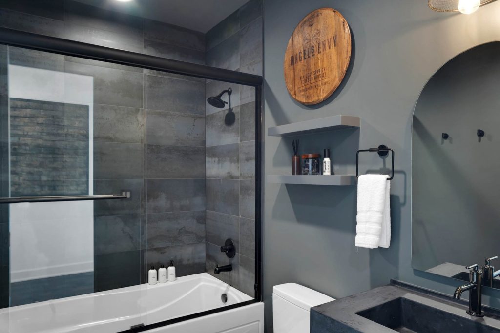 Modern bathroom with gray tiles, glass shower, white bathtub, minimalist shelves, towels, and wooden wall decor. Sleek and calm.