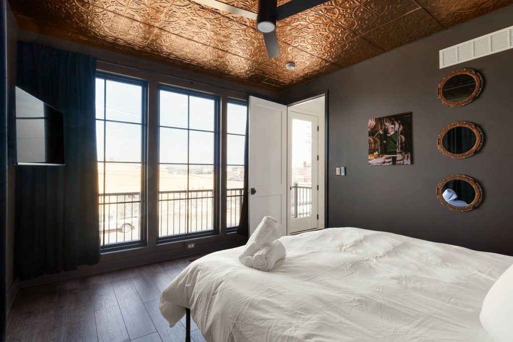 Cozy modern bedroom with white bed, dark walls, large windows, and ornate copper ceiling. Warm, inviting ambiance.