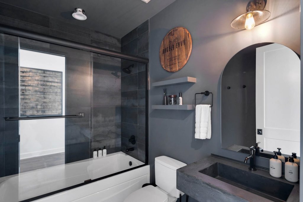 Optimized Alt Text: Modern bathroom with dark gray walls, glass-enclosed tub, circular wall art, sleek shelves, and concrete sink with rectangular mirror. Character Count (Alt Text): 124 Image Title: Contemporary Modern Bathroom with Dark Gray Walls, Glass-Enclosed Bathtub, Concrete Sink, and Sleek Shelving