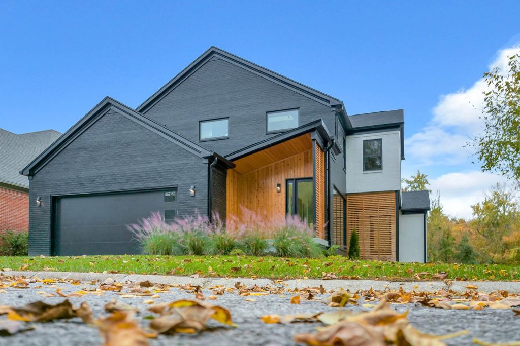 Modern house with dark gray brick, large windows, wooden entry, autumn leaves, and green foliage under a clear blue sky.
