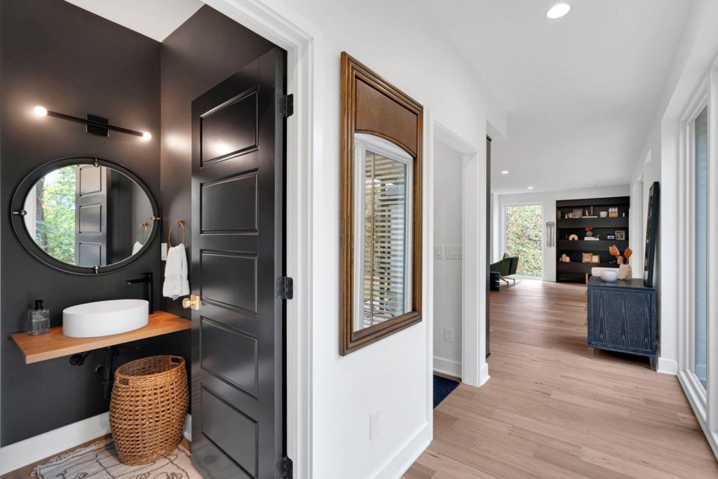 Modern hallway with light wood floors, white walls, a dark bathroom doorway with round mirror, and large framed wall mirror.