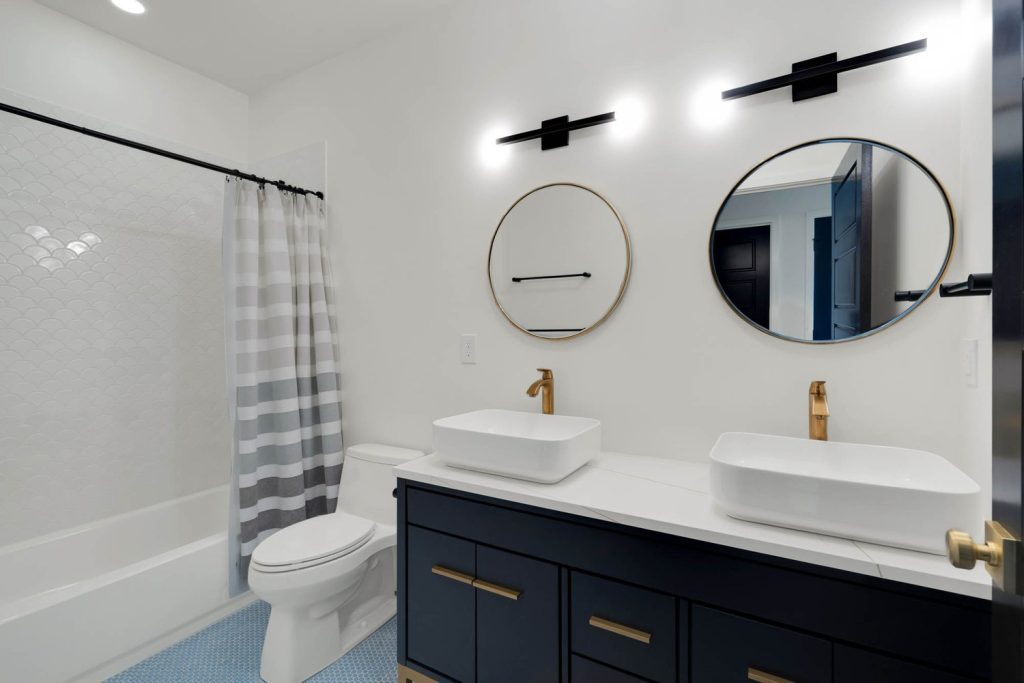 White bathroom with a blue vanity, twin round mirrors, gold faucets, vessel sinks, and a gray striped shower curtain.