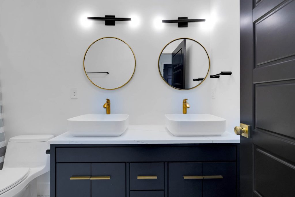 Bathroom vanity with two round mirrors, white vessel sinks, gold faucets, and dark cabinetry set against crisp white walls.