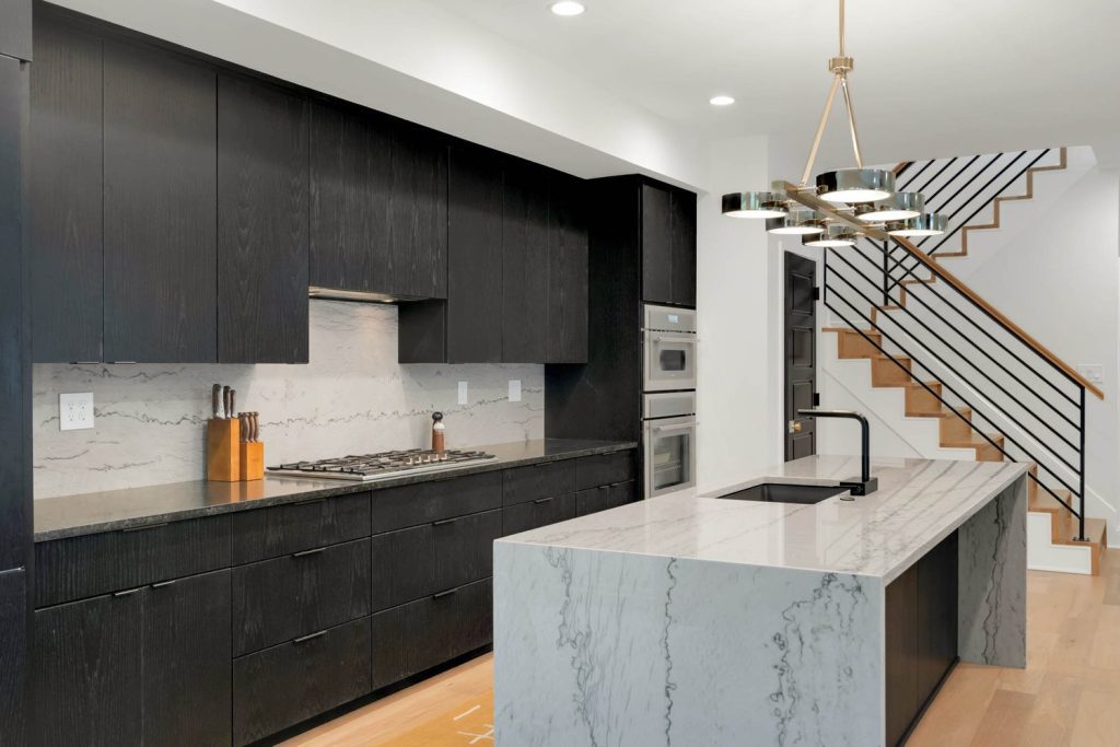 Modern kitchen with black cabinets, marble island, sleek faucet, and chandelier; staircase visible in background.