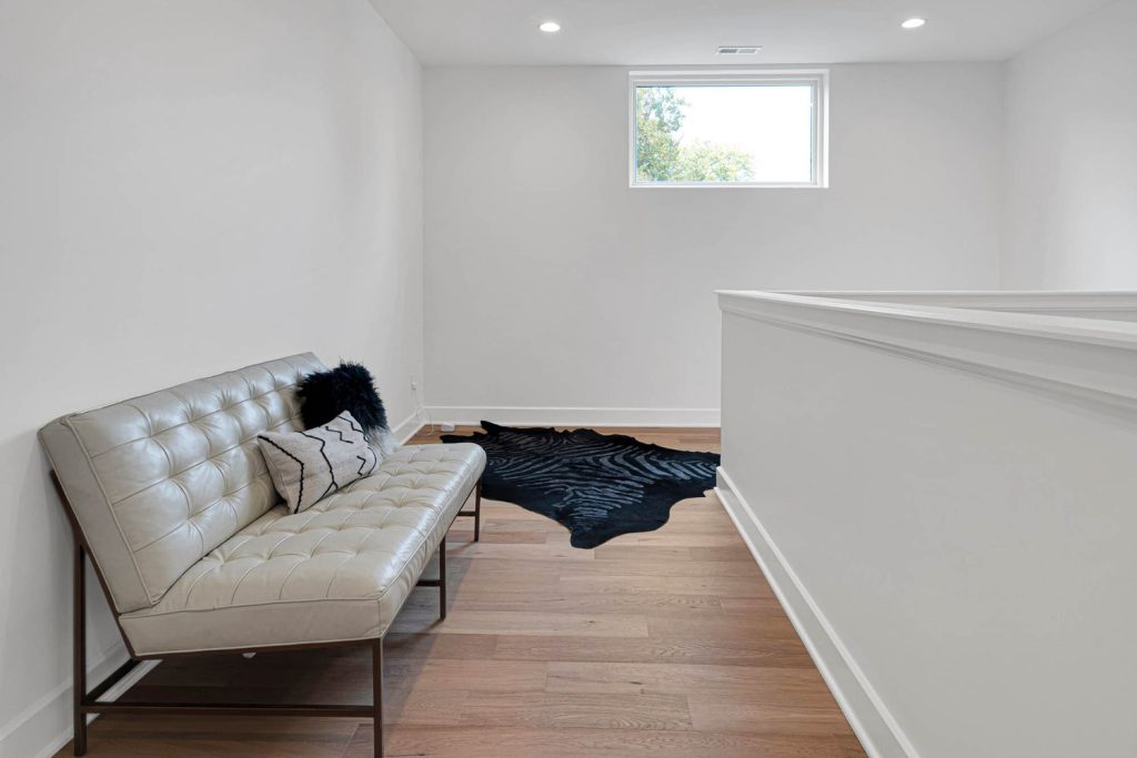 Minimalist room with beige leather sofa, black cushions, dark rug, and wood floors, softly lit by a small window. Image Title (under 120 characters, different from alt text): Contemporary sitting area with neutral tones and clean lines, styled for a custom home interior with subtle natural lighting