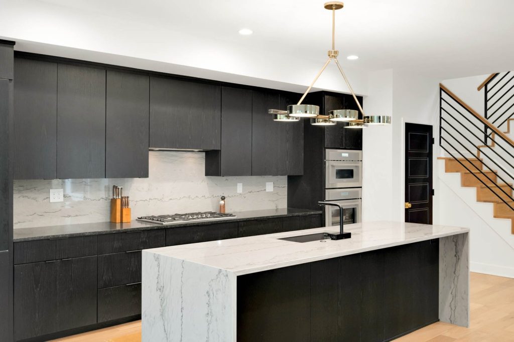 Sleek modern kitchen with dark cabinets, marble island, gold pendant light, and built-in oven and stove.