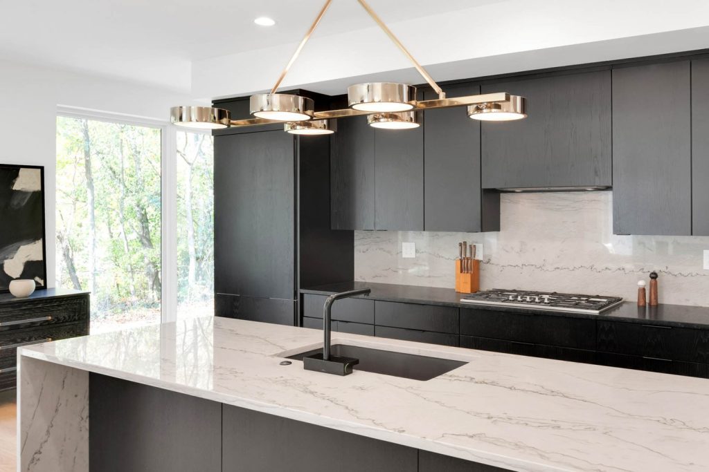 Modern kitchen with black cabinets, marble countertop, and gold linear chandelier. Natural light brightens the space.