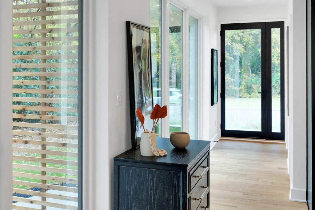 Bright modern entryway with large windows, black door, dark wooden cabinet, vase of orange leaves, and round beige bowl.