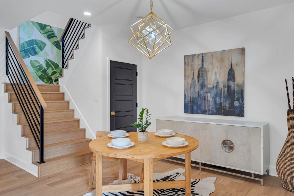 Modern dining area with circular wooden table for four, geometric chandelier, abstract wall art, and leafy wallpapered stairs.