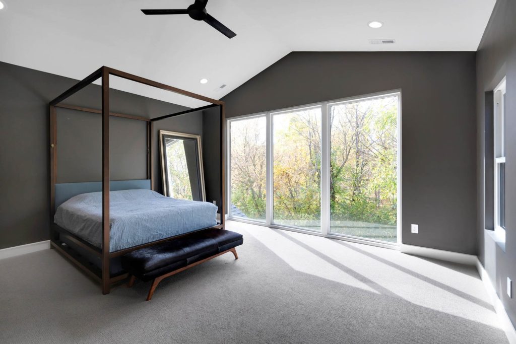 Four-poster bed with blue bedding faces large windows showing autumn trees. A leaning mirror and sleek ceiling fan add clean style.
