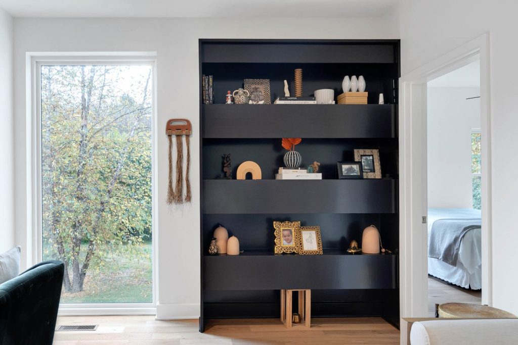 Modern black bookshelf with framed photos, vases, and art; large window reveals lush green garden. Stylish, serene room.