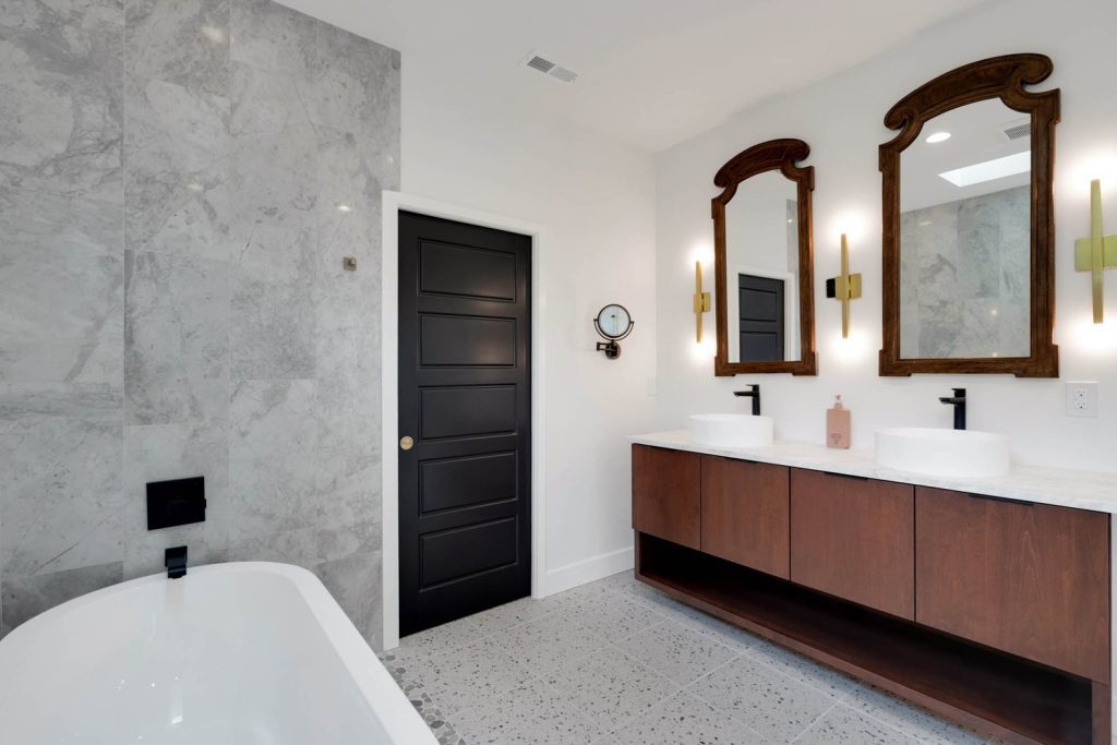 Freestanding tub beside a wood vanity with dual sinks, ornate mirrors, and a black door, creating a refined spa-like bathroom.