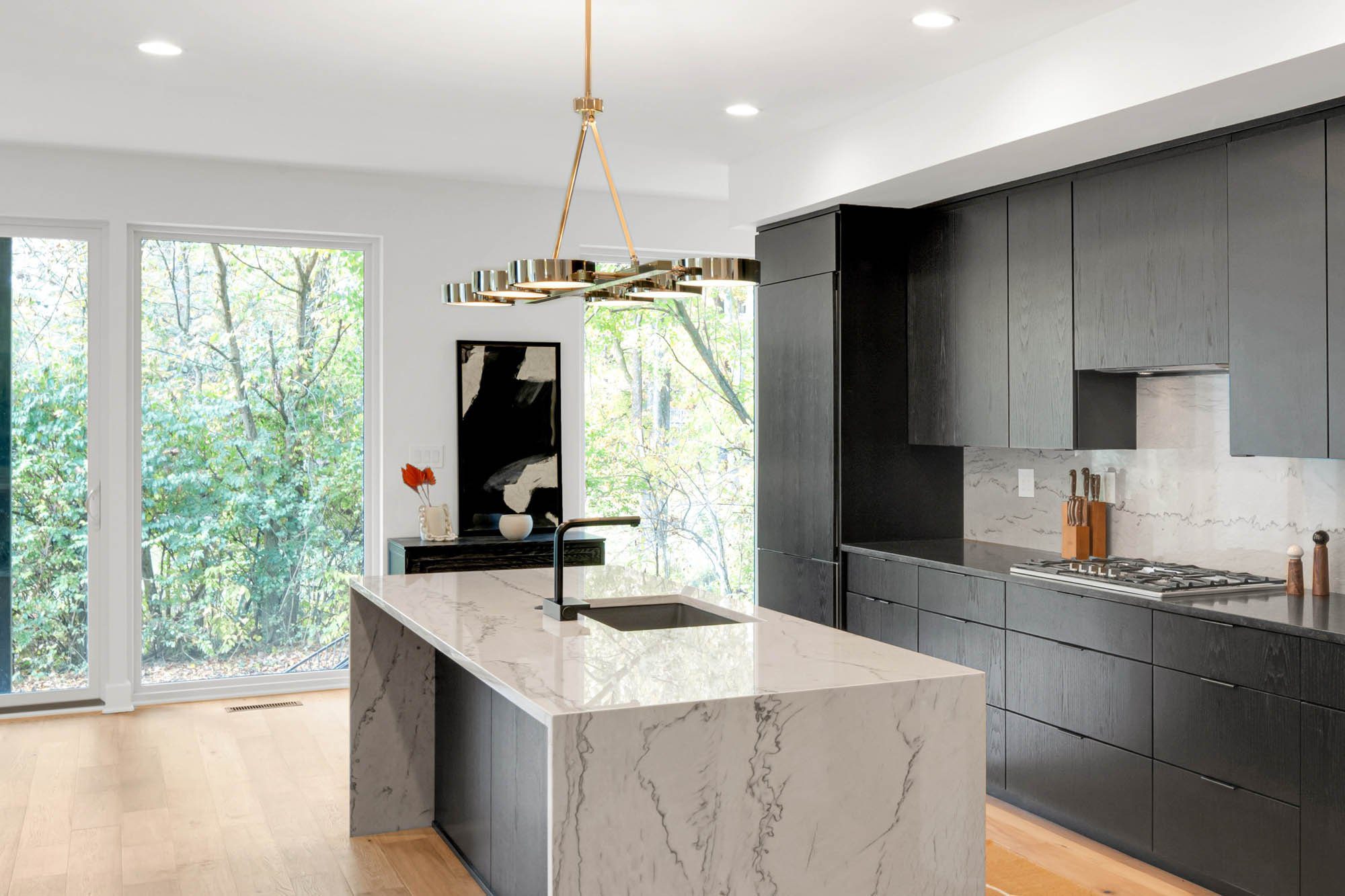 Modern kitchen with marble island, black cabinets, gold pendant light, and large windows overlooking trees.