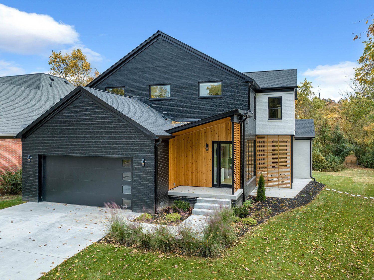 Modern home exterior with black siding, wood accents, large windows, gray driveway, and lush lawn under blue sky.