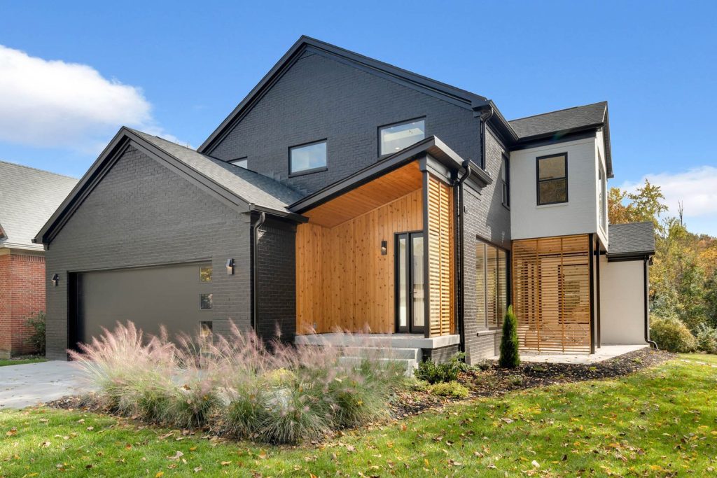 Modern two-story house with dark gray brick, wood accents, large windows, sloped roof, and landscaped yard.