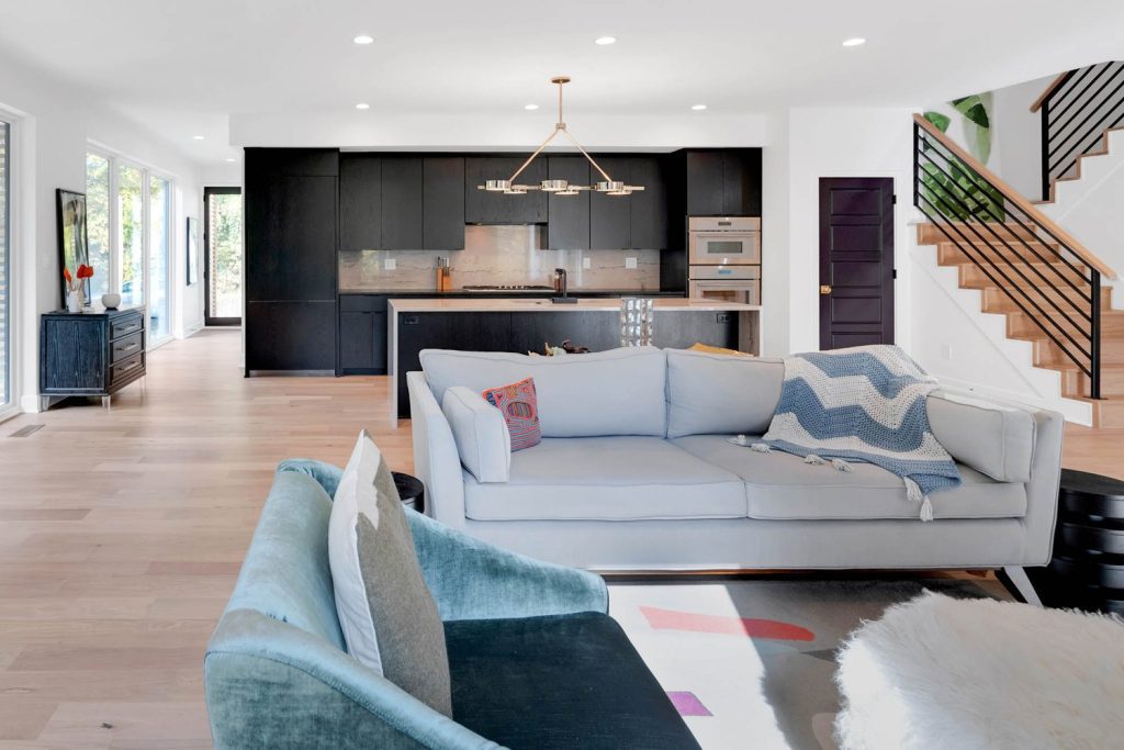 Modern living room with light blue sofas, colorful pillows, chevron throw, and sleek black kitchen in an open-plan design.
