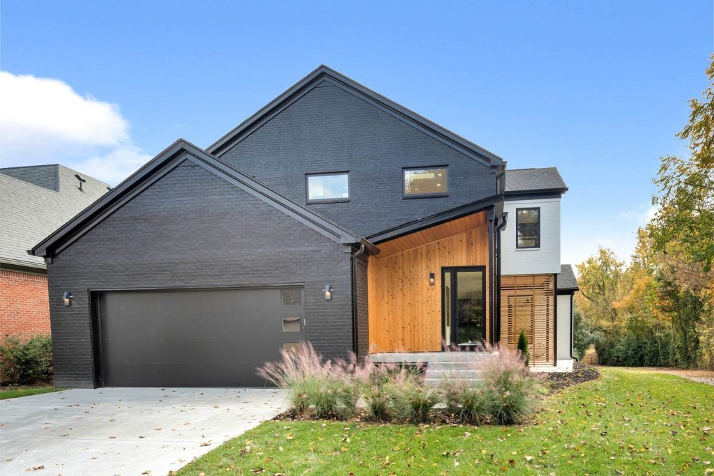 Modern house with dark gray siding, wooden accents, large windows, slanted roof, and landscaped lawn with ornamental grasses.