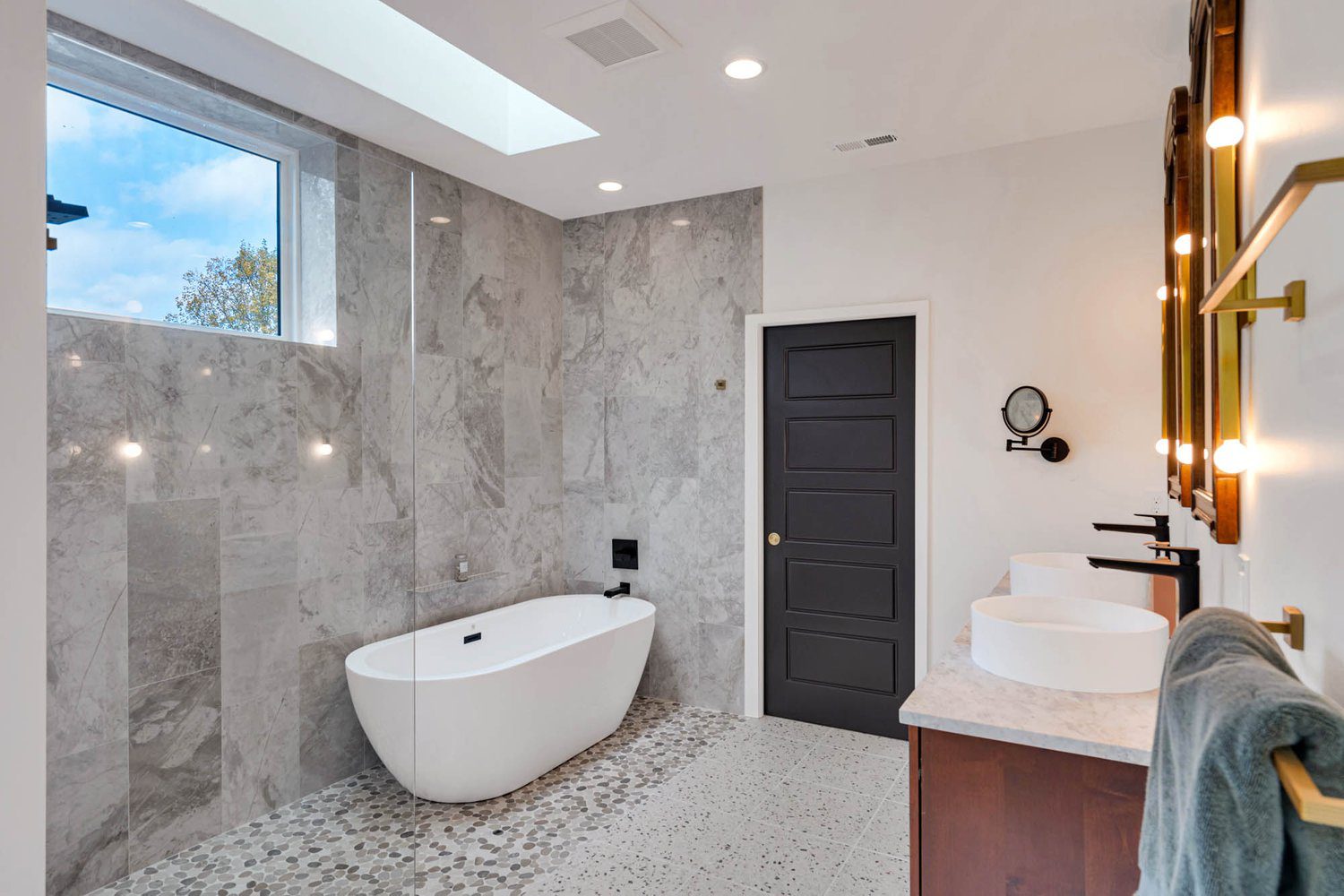 Modern bathroom with gray marble walls, freestanding tub, wooden vanity with double sinks, and skylight.