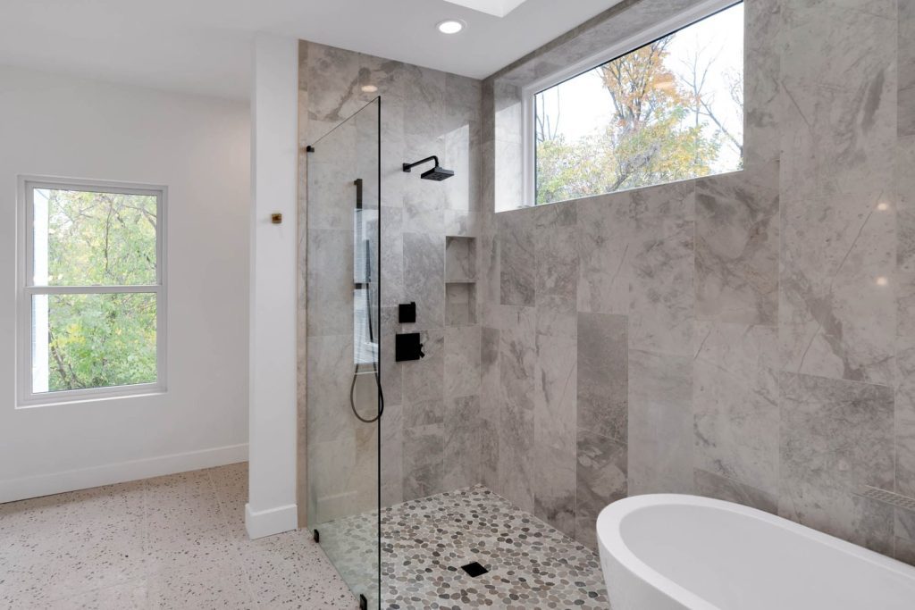 Bathroom with glass shower, gray marble tiles, and pebble floor. Sunlight from window creates a calm, spa-like vibe.