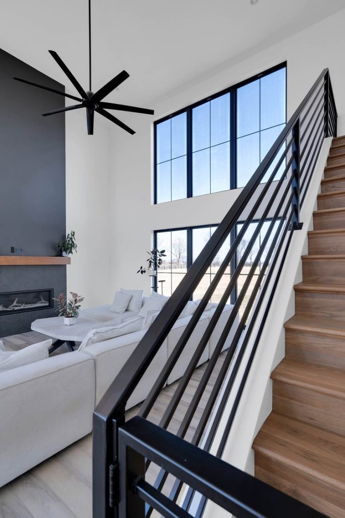 Louisville modern living room with high ceilings, white sectional, wall-mounted fireplace, wooden staircase, and black ceiling fan.