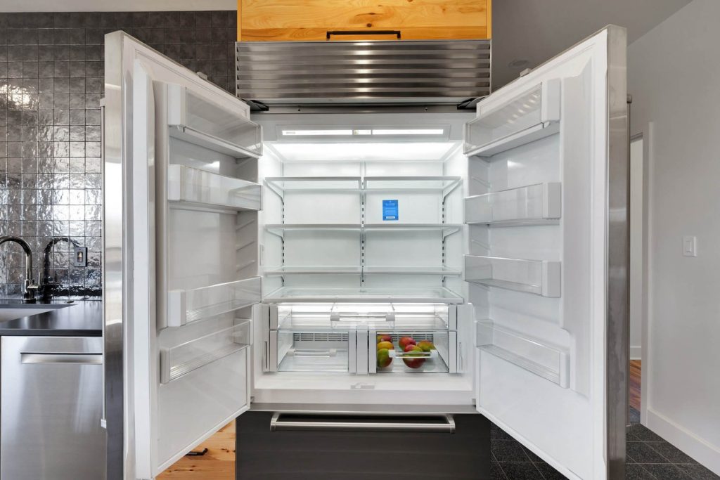 Open double-door refrigerator with few apples, bright lighting, and modern stainless steel accents in a sleek kitchen