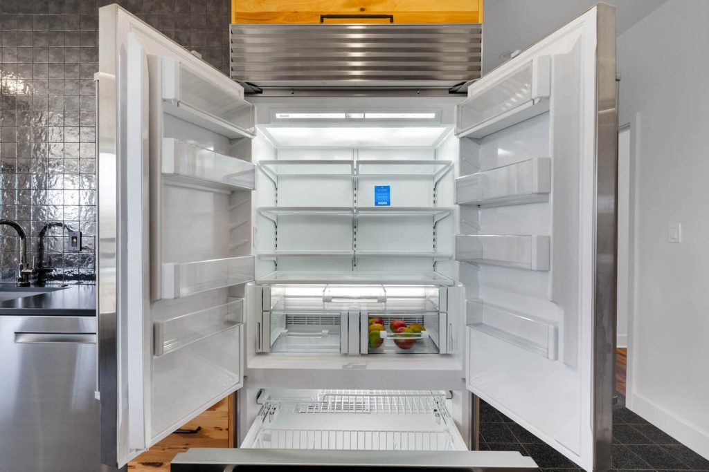 Open commercial-style refrigerator in a modern kitchen with clean shelves and a few apples in the bottom drawer