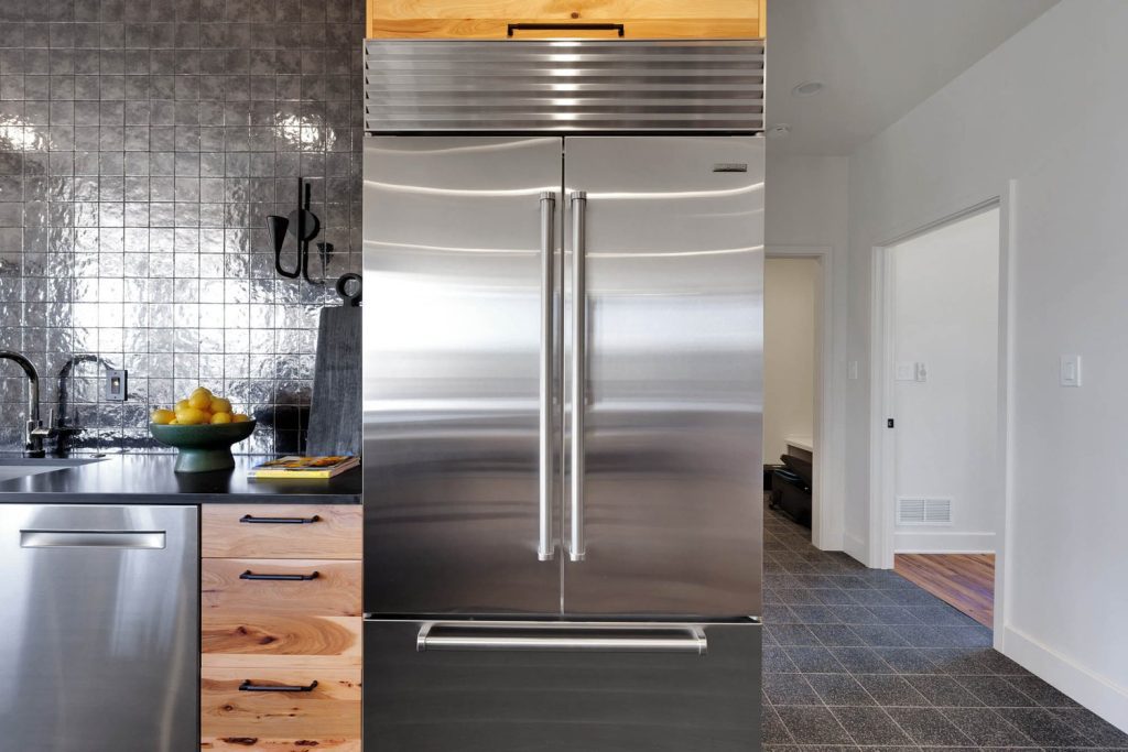 Stainless steel double-door fridge in a modern kitchen with wooden cabinets and reflective tile, sleek and contemporary design