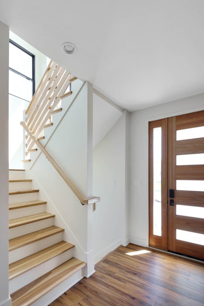 Modern entryway with light wood staircase, clean railing, and a sleek wooden door with horizontal glass panels