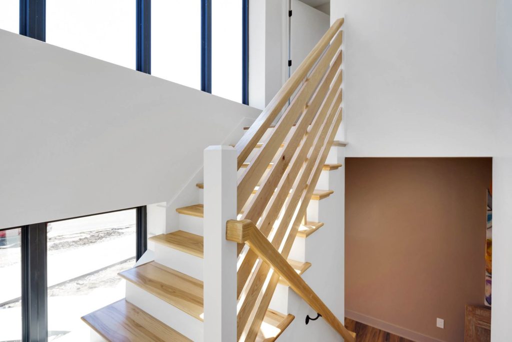 Modern wooden staircase with light-toned steps, horizontal railing, and large windows bringing in natural light