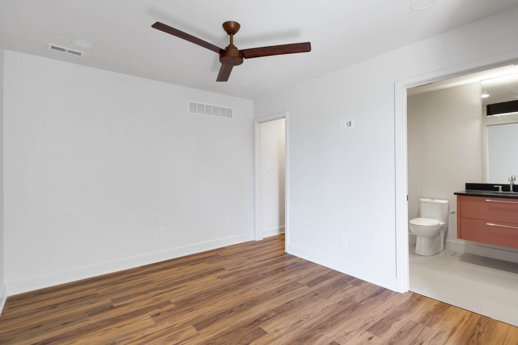 Empty modern room with white walls, wood flooring, and ceiling fan, opening to a bathroom, showcasing new home construction design ideas