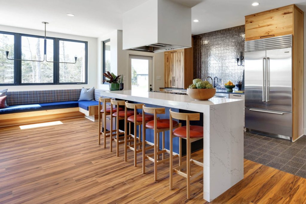 Modern kitchen with marble island, wooden floors, red-cushioned stools, blue corner seating, and large bright windows