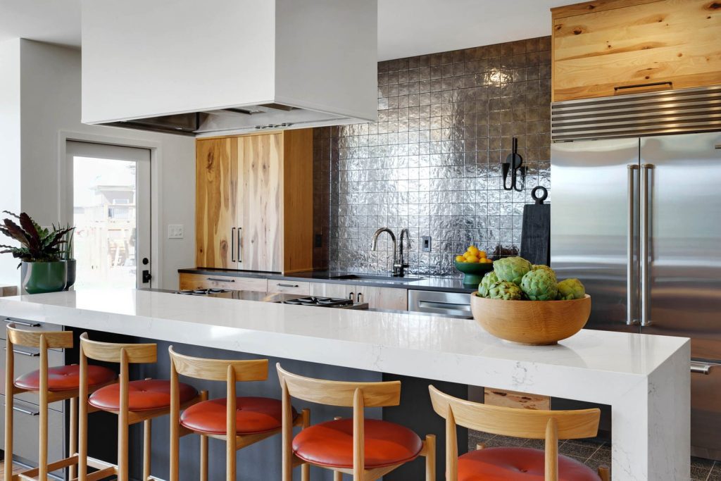 Modern kitchen with white countertop, wooden bar stools with red cushions, metallic backsplash, and wooden cabinets
