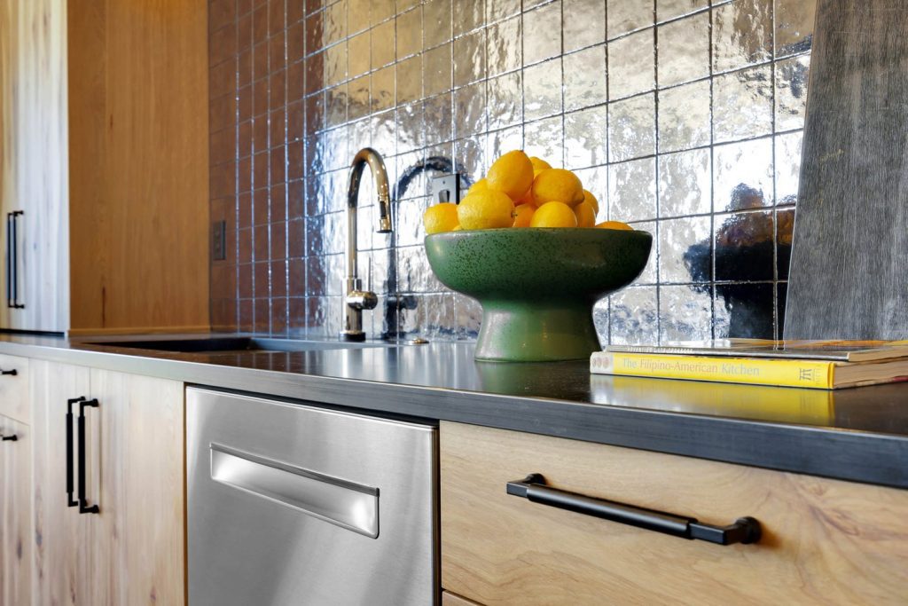 Modern kitchen with glossy tile backsplash, stainless steel sink and dishwasher, and green bowl of lemons on black countertop