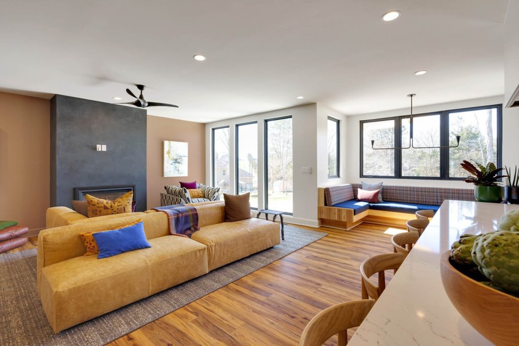 Modern living room with tan sectional, blue and brown pillows, large windows, wood floors, bar stools, and window nook