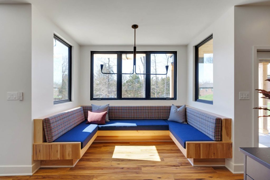 Cozy corner nook with blue cushioned seating, plaid backrests, accent pillows, and large windows with soft natural light