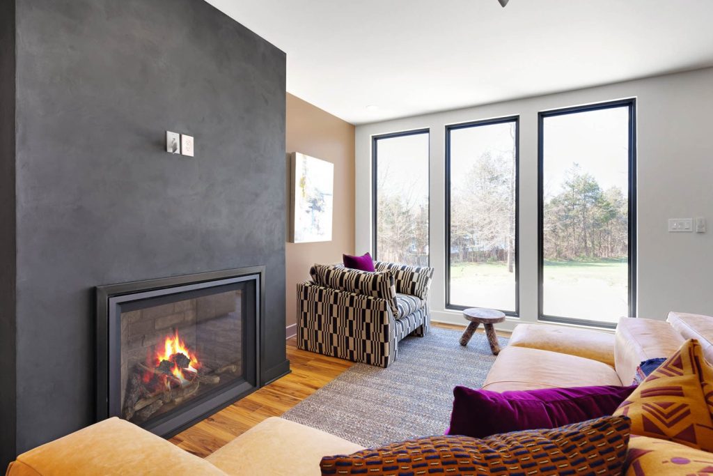 Modern living room with dark gray fireplace wall, striped armchair, and large windows overlooking a sunny garden
