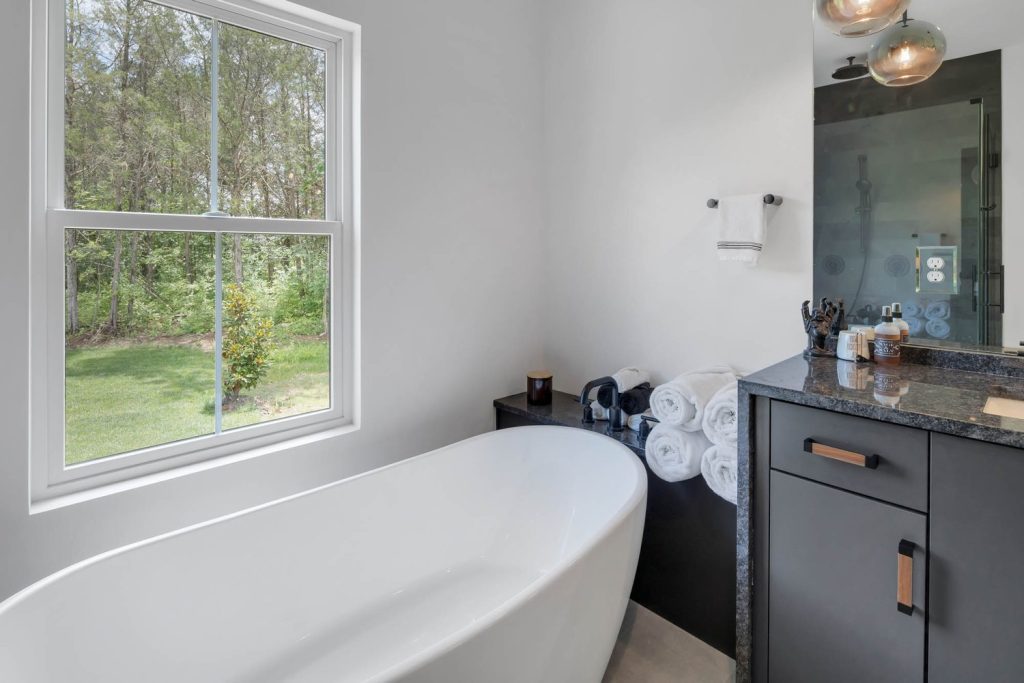 Bathroom with freestanding tub by window, gray dual-sink vanity, glass shower, and pendant lights in a sleek modern design