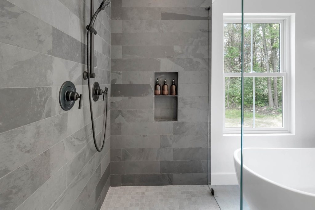 Bathroom with gray tiled walls, chrome shower fixtures, built-in shelf with toiletries, and a window overlooking greenery