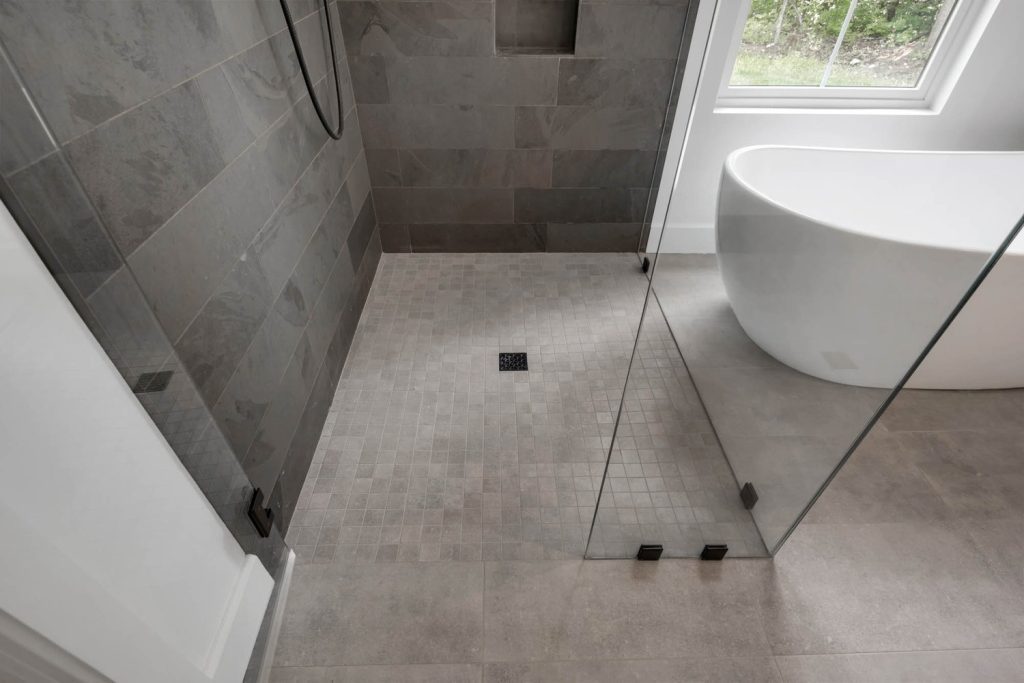 Minimalist bathroom with walk-in glass shower, gray tiles, and white freestanding tub by a window with natural light