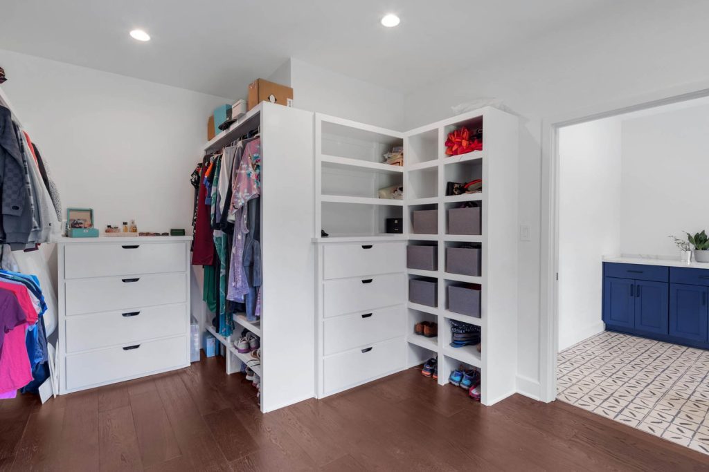 Spacious walk-in closet with white shelves, organized clothes, shoes, boxes, warm wood floor, and bright overhead lighting