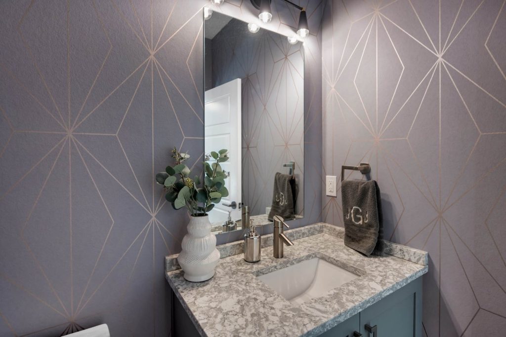 Bathroom with geometric wallpaper, lit vanity mirror, patterned countertop, and white vase with greenery for a fresh look