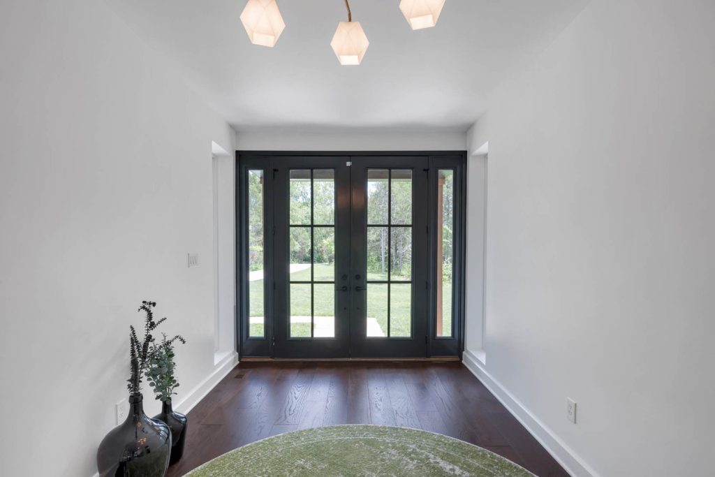 Entryway with dark double doors opening to a green lawn, white walls, wood floors, ceiling lights, and a soft green rug.