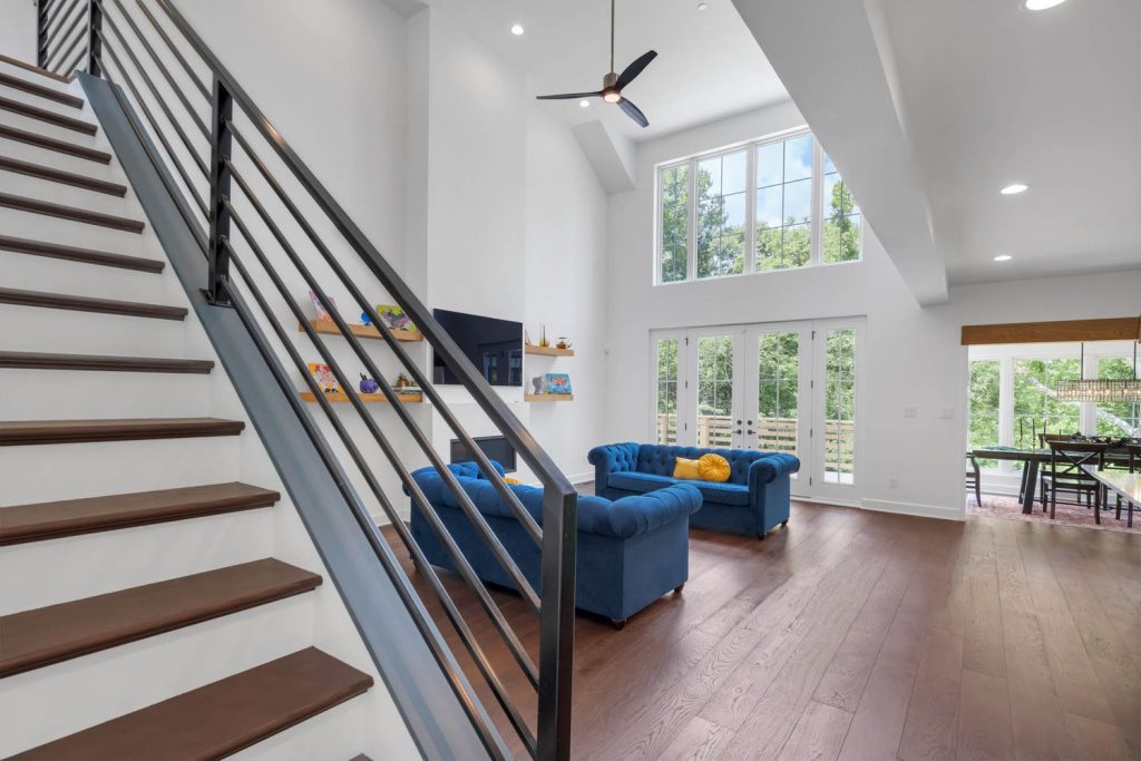 Spacious living room with tall windows, blue sofas, ceiling fan, and wood floors filled with soft natural daylight.