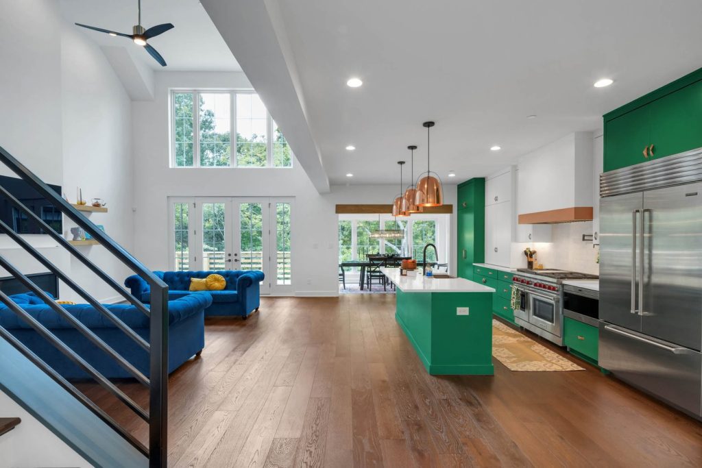 Open kitchen and living space with green cabinets, copper pendants, blue sofa, and tall windows overlooking the garden.