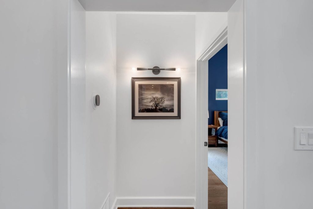 Narrow hallway with white walls, black-and-white landscape photo under a wall lamp, and a peek of a blue-painted room